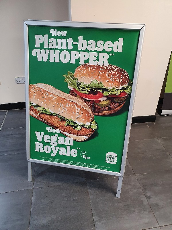 A green poster advertising Burger King's new plant-based options, featuring images of a Plant-based Whopper and a Vegan Royale. The text highlights these new offerings with the Burger King logo at the bottom right corner.