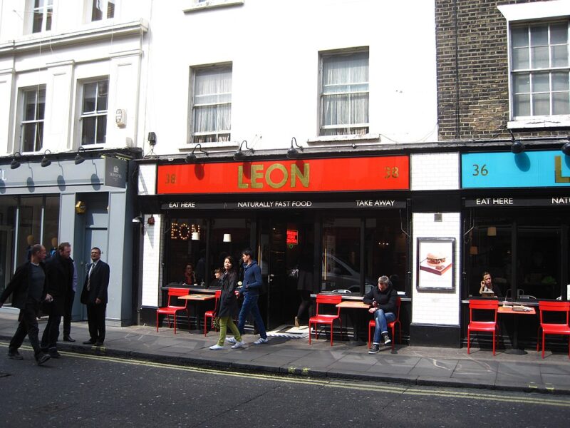 Street view of Leon Restaurant on Old Compton Street, London. The restaurant has a red and orange sign, with outdoor seating where a few people are sitting and others walking by.