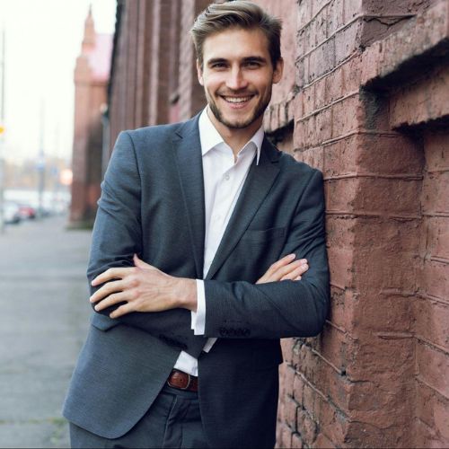 A man in a grey suit standing against a brick wall outside, smiling and crossing his arms.