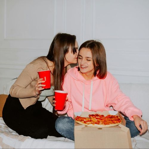 Two women sitting on a bed, sharing a pizza and drinking from red cups. One woman is whispering into the other's ear, making her smile.