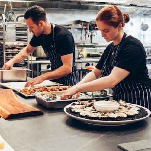 Two chefs in a professional kitchen, diligently preparing food on trays, surrounded by culinary equipment and ingredients.