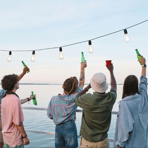 A group of friends standing by a lakeside at sunset, raising their drinks in a toast under string lights.