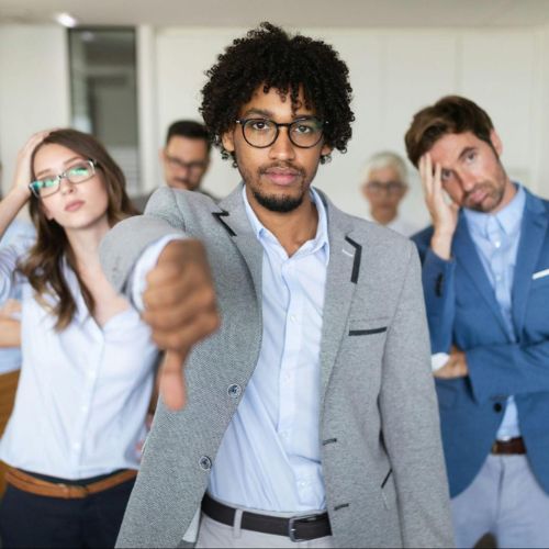 A group of businesspeople gathered together, with one person in the foreground giving a thumbs-down gesture, while others look stressed or disappointed.