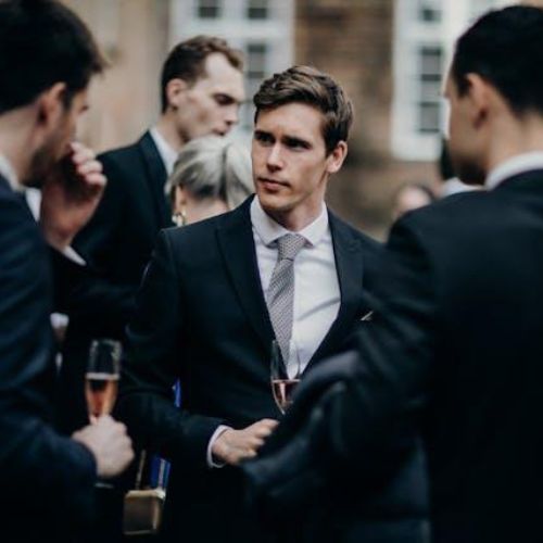 A man dressed in a suit standing among a group of people at a formal event, holding a glass of wine and engaging in conversation.