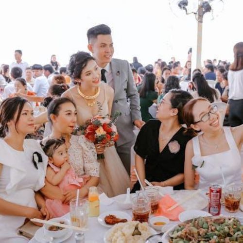 A wedding reception scene with a bride and groom standing and smiling among a crowded table of guests. The bride is holding a bouquet, and everyone is enjoying the celebration.