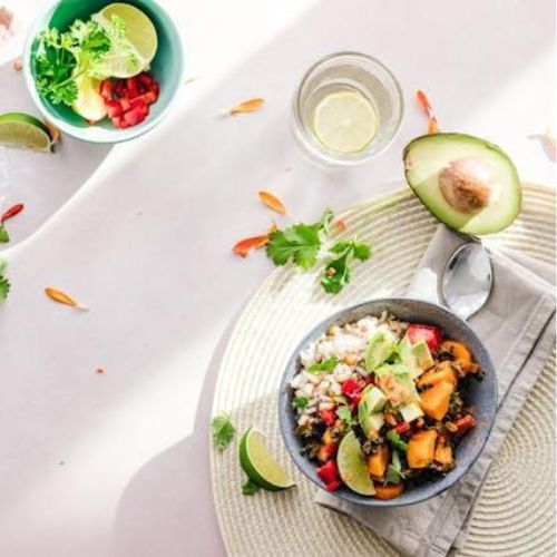 A healthy breakfast bowl filled with a colorful mixture of fruits, vegetables, and grains, placed on a woven placemat. Nearby are a glass of water with a slice of lemon, half an avocado, and a small bowl containing lime slices, parsley, and diced tomatoes.