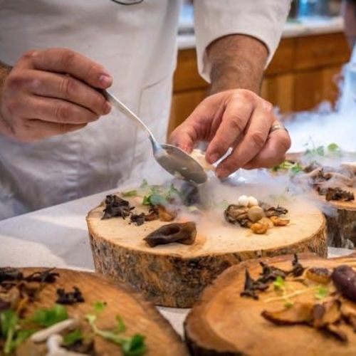 A chef's hands artfully plating a dish on rustic wooden serving boards. The dish includes a variety of mushrooms, vegetables, and garnishes