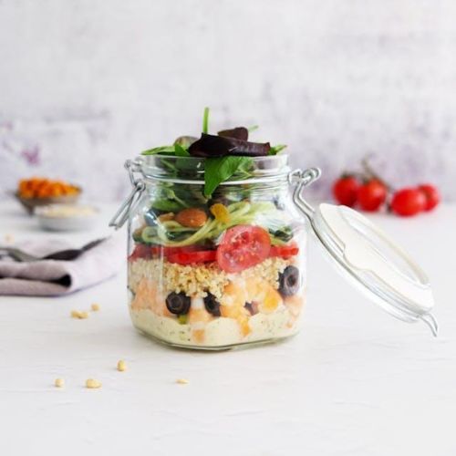 A jar filled with a layered salad, including ingredients such as cherry tomatoes, olives, greens, and grains, set on a white countertop.