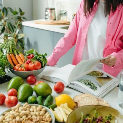 A person in a pink shirt is flipping through a cookbook on a kitchen counter filled with various fresh vegetables, fruits, and nuts.