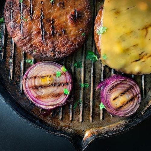 A close-up of a grill with two burger patties, one with melted cheese, and two red onion slices, all garnished with herbs.