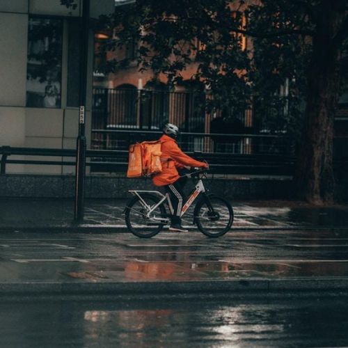 A person in an orange jacket riding a bicycle with a large orange delivery backpack, traveling in a city area at night with wet roads reflecting lights.