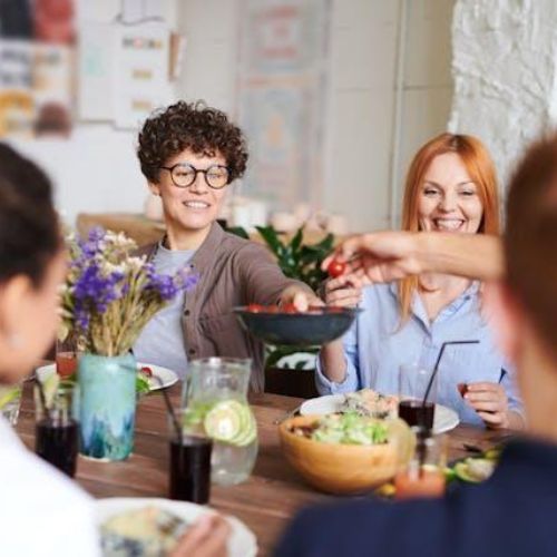 A group of people sitting around a wooden dining table, sharing a meal. There are plates of food and drinks on the table, along with a vase of flowers. One person is passing a bowl of food to another. Everyone appears to be smiling and enjoying the meal together.