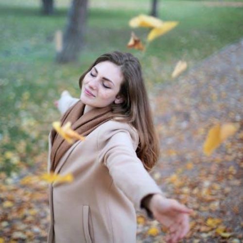 A woman in a beige coat enjoying a stroll in a park during autumn. She has her arms outstretched and is smiling with her eyes closed as yellow leaves fall around her.