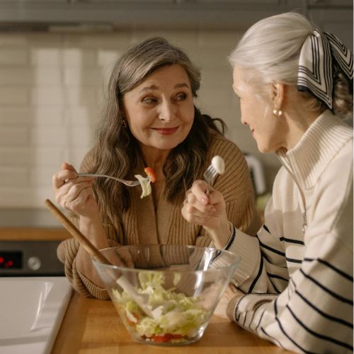 Two elderly women sitting in a kitchen, sharing a bowl of salad and smiling at each other. One is holding a fork with salad, and the other is gesturing while talking.