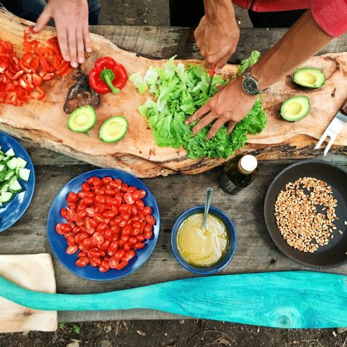 A top view of hands preparing a meal on a rustic wooden table with various ingredients, including chopped red peppers, avocado halves, lettuce, cherry tomatoes, and pine nuts. There is also a bowl of dressing and a large wooden cutting board.