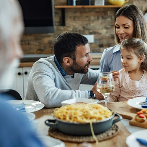 A family of three, two adults and a child, sitting at a dining table, enjoying a meal together. There is a pan of pasta on the table, and they appear to be smiling and conversing.