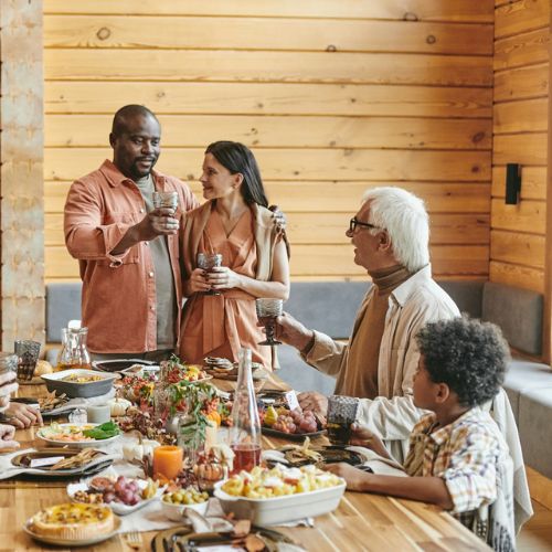 A group of people gathered around a large dining table set with various dishes. They are raising their glasses in a toast inside a cozy, wooden-themed room.