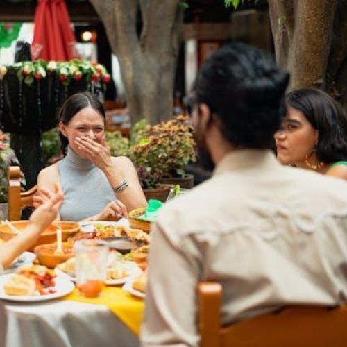 A group of people sitting at an outdoor restaurant table, eating and laughing. One person has their hand over their mouth while smiling, engaged in a joyful conversation.