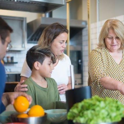 A group of people of different ages in a kitchen, working together to prepare food. They are surrounded by various bowls and ingredients, and one person is holding an orange.