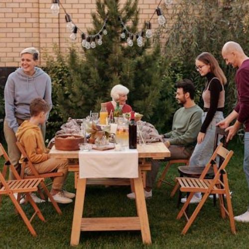 A family gathering in a backyard around a table set for a meal. There are lights strung up above, and everyone is engaging in conversation and preparing to eat.