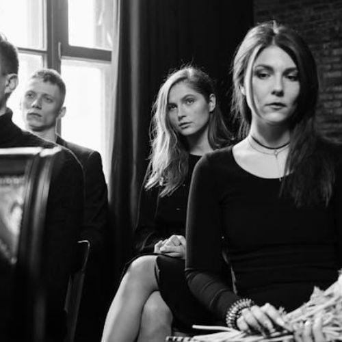 A group of people sitting in a room with a somber atmosphere. They appear to be attending a formal or serious event, dressed in dark clothing, with expressions reflecting contemplation.