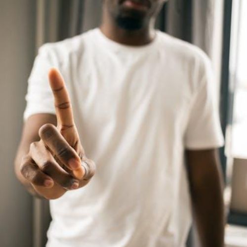 A person in a white t-shirt extending their index finger towards the camera against an indoor background.