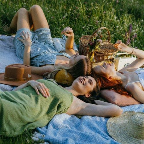 Three people lying on a blanket in a grassy field, enjoying a sunny day with a picnic basket nearby. They appear relaxed and happy.