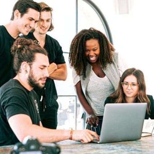 A group of five people, gathered around a laptop on a table, smiling and discussing something on the screen. There are notebooks, a camera, and drinks on the table.