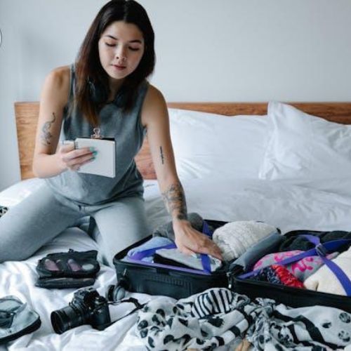 A woman sitting on a bed, packing a suitcase. They are holding a notepad and pen, with various items such as clothes, a camera, and accessories around them.