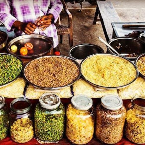 A display of various grains and pulses in bowls and jars at a market stall, with a person standing behind the counter.