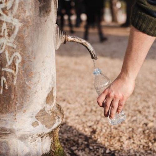 A person filling a plastic bottle with water from an old outdoor faucet.