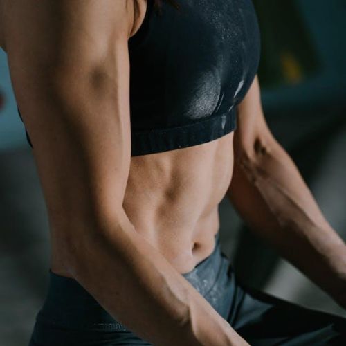 A close-up of a woman's upper body in workout attire. She is wearing a black sports bra and dark leggings, showcasing her muscular arms and toned abs