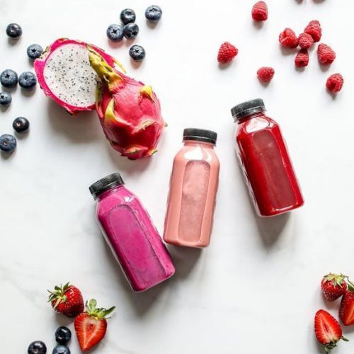 An overhead view of three bottles of homemade fruit smoothies arranged on a white surface. Surrounding the bottles are fresh berries and a sliced open dragon fruit