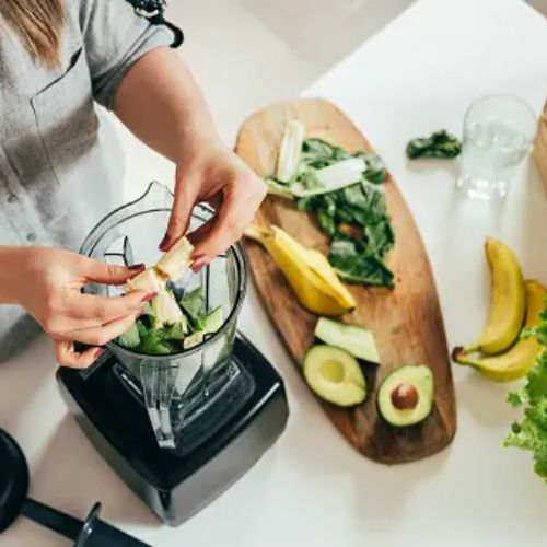 A woman preparing a smoothie in a blender in a kitchen. She is adding fresh ingredients, such as avocados, bananas, and leafy greens, to the blender