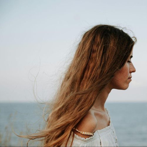 A profile view of a person with long hair standing near the ocean, facing sideways. The background features the sea and a clear sky.