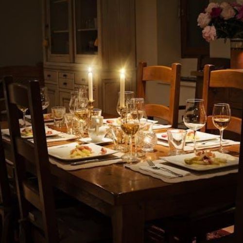 A warmly lit dining table setup with six chairs around it. There are lit candles, plates of food, wine glasses, and tableware arranged neatly.