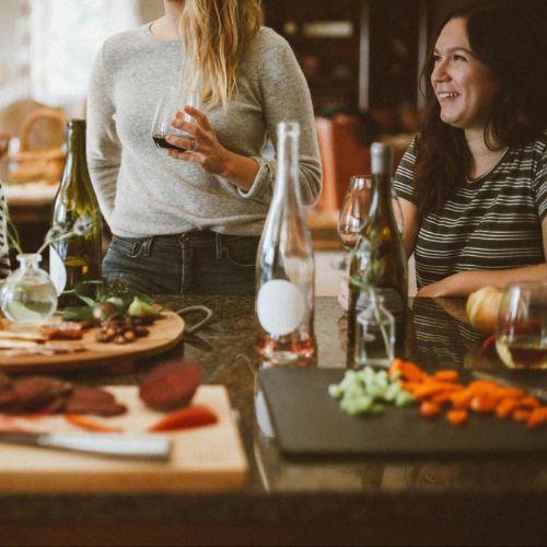 A group of people standing and chatting around a kitchen island with wine glasses, bottles of wine, and cutting boards with various food items placed on it.