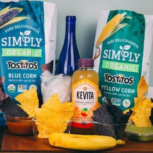 A display of food products on a table, including bags of Simply Organic Tostitos chips, a bottle of Kevita sparkling probiotic drink, a blue glass bottle, salsa, guacamole, and a banana.