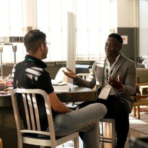 Two men in conversation while seated at a table in a well-lit indoor space furnished with chairs and tables, possibly an office or conference setting.