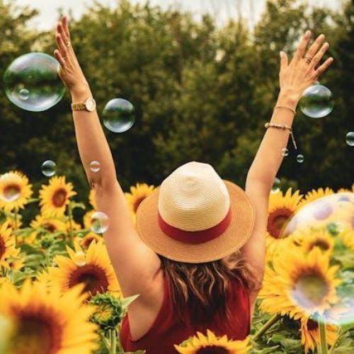 A woman standing in a field of sunflowers with their arms raised, surrounded by floating bubbles. They are wearing a straw hat and a red outfit.