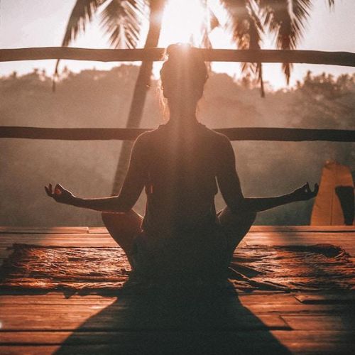 A woman sitting cross-legged on a wooden deck in a meditative pose during sunrise or sunset, with palm trees and a forested area in the background.