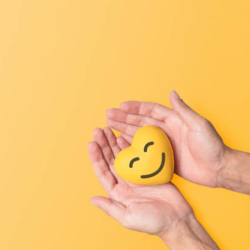 Two hands holding a yellow emoji stress ball with a smile, set against a plain yellow background, representing care or positivity.