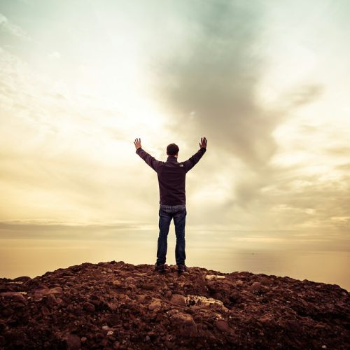 A person stands on mountain peack, high above a lake under a cloudy sky, symbolizing achievement and solitude.