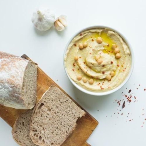 An overhead view of a loaf of rustic bread partially sliced on a wooden cutting board next to a bowl of creamy hummus. The hummus is garnished with olive oil, whole chickpeas, and red pepper flakes