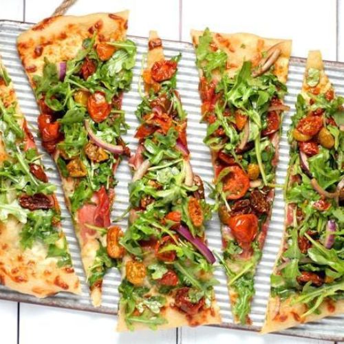 An overhead view of a rectangular pizza cut into strips. The pizza is topped with arugula, roasted cherry tomatoes, red onions, and cheese