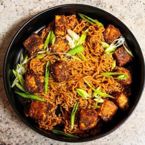 A bowl of spicy noodles with crispy tofu and green onions, garnished with sesame seeds