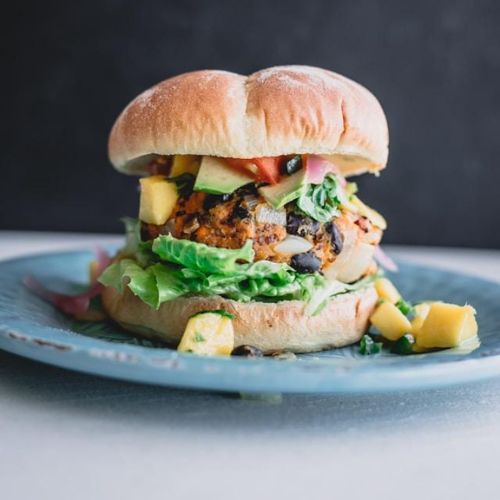 A close-up of a veggie burger with various toppings, including lettuce, cheese, avocado slices, and diced mango.