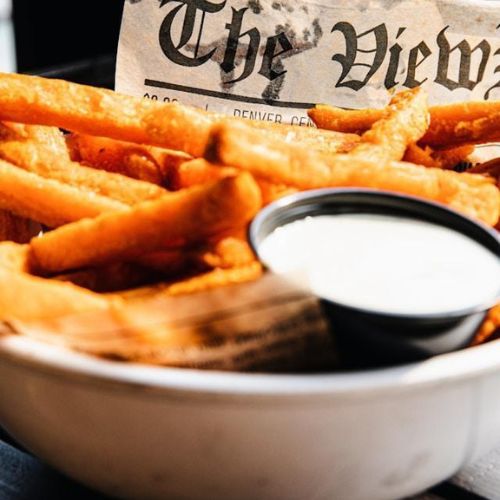 A close-up of a bowl of golden crispy French fries, lined with a piece of newspaper with text visible. A small container of dipping sauce is placed next to the fries