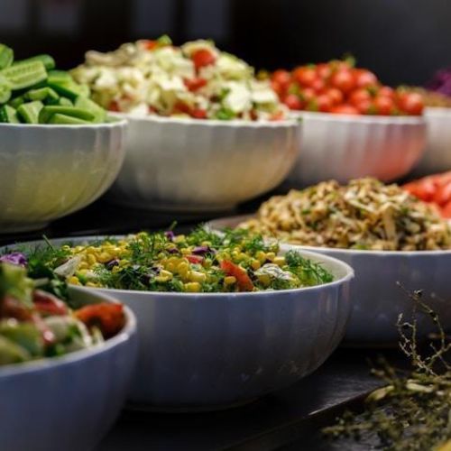 A colorful array of salads and fresh ingredients displayed in white bowls. The bowls contain various chopped vegetables, grains, and greens, including cucumbers, tomatoes, and leafy greens