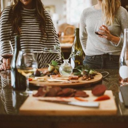 A group of women gathered around a kitchen island, enjoying wine and snacks. The table is set with various appetizers, such as sliced vegetables, meats, and cheeses, along with wine glasses and bottles